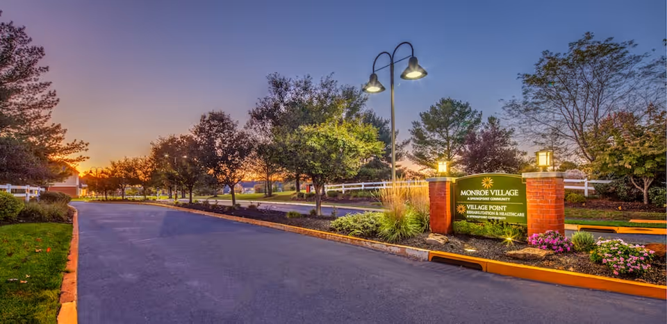 Landscaped driveway entrance at sunset showing a lit brick sign for Village Point Rehabilitation & Healthcare with trees and street lamps.