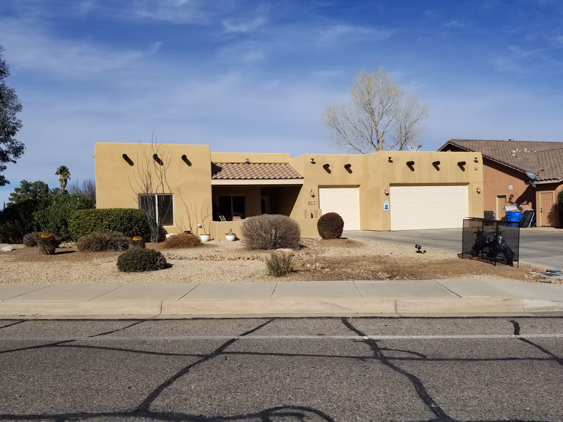 Front exterior view of a single-story building with a beige stucco finish and a tiled roof. The building has two white garage doors, a small covered porch area, and desert landscaping with bushes, small trees, and gravel. The sky is clear with some clouds.