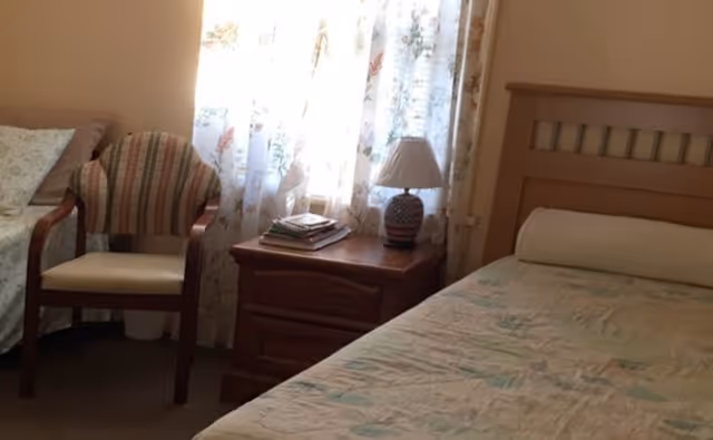 A cozy bedroom in a senior facility with a bed, chair, nightstand, lamp, and floral curtains.