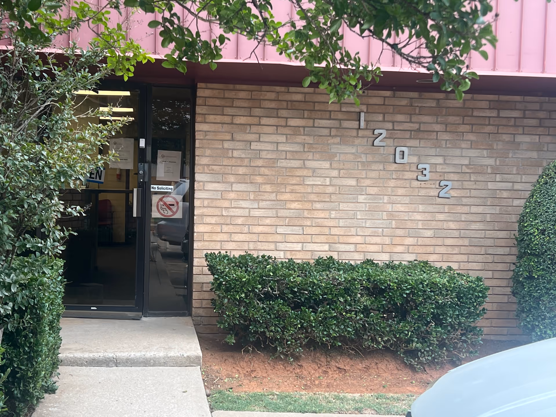 Entrance to a brick facility with a glass door, greenery, and a pink awning.