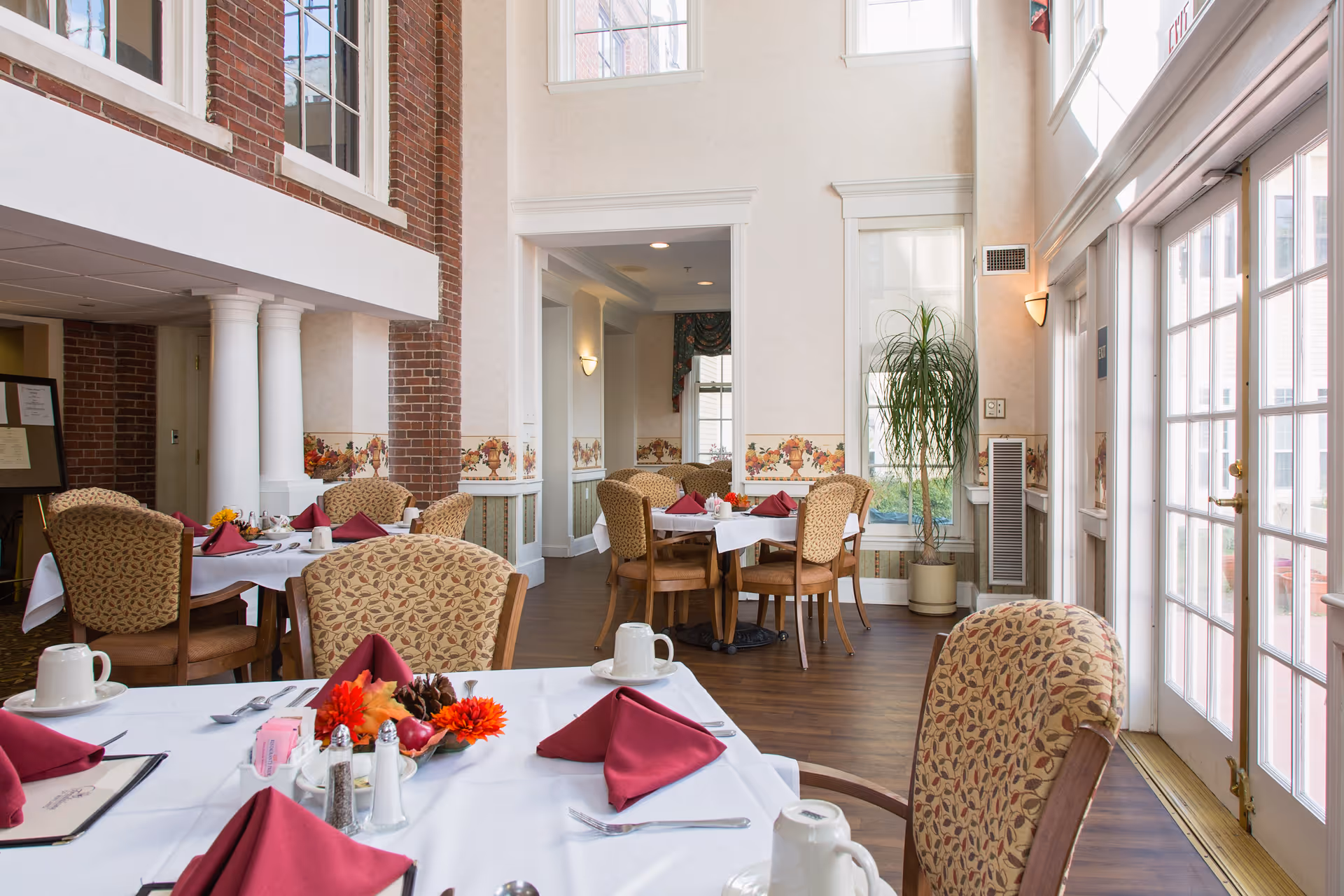 A bright dining room with tables set for a meal, featuring white tablecloths, burgundy napkins, and floral centerpieces. The room has large windows and glass doors letting in natural light, patterned upholstered chairs, brick and cream-colored walls with decorative trim, and a tall potted plant near the window.