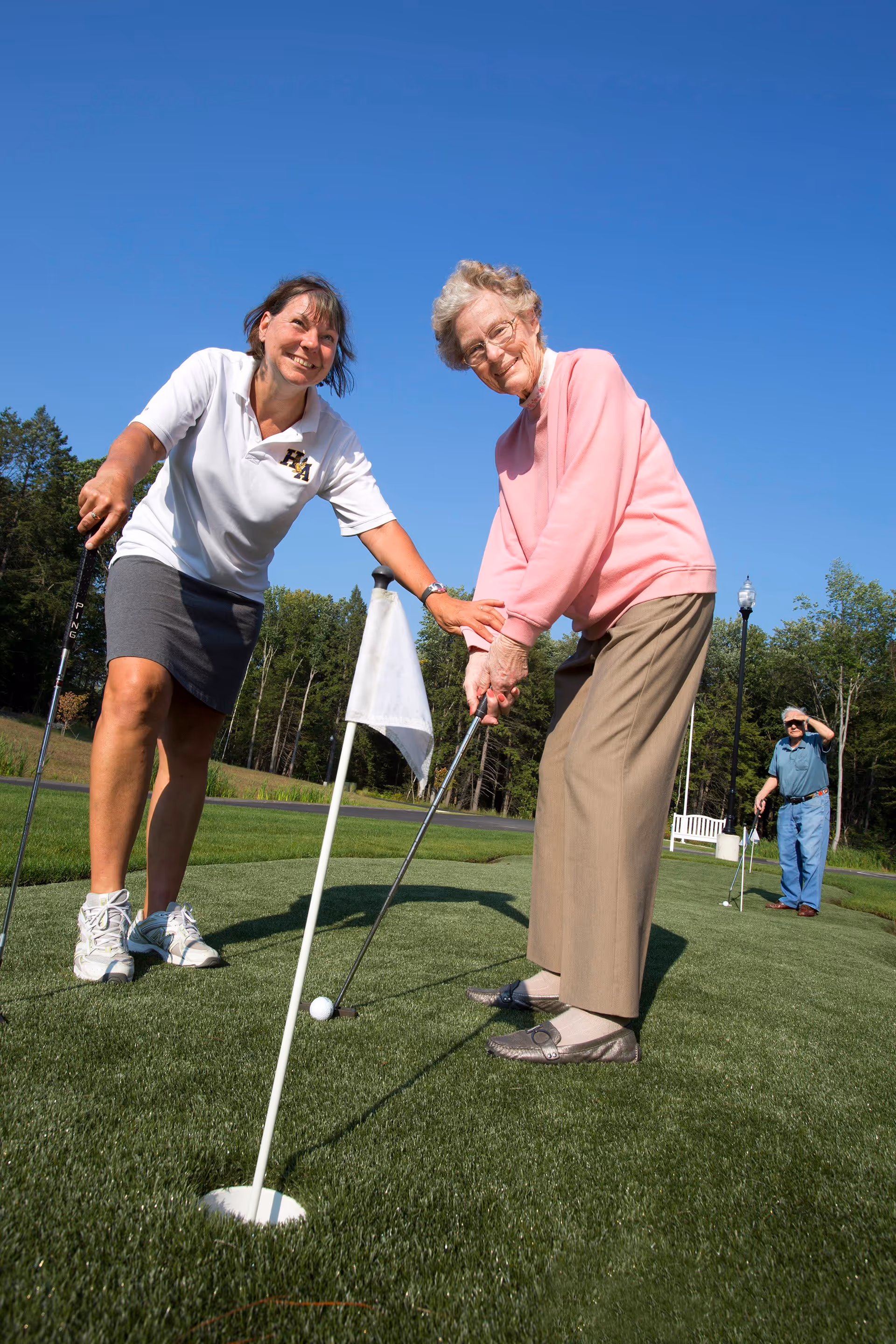 An elderly woman in a pink sweater and beige pants is putting a golf ball near a hole on a putting green, assisted by a younger woman in a white polo shirt and gray skirt. Another elderly man stands in the background on the green, shielding his eyes from the sun. The scene is outdoors on a sunny day with a clear blue sky and trees in the background.