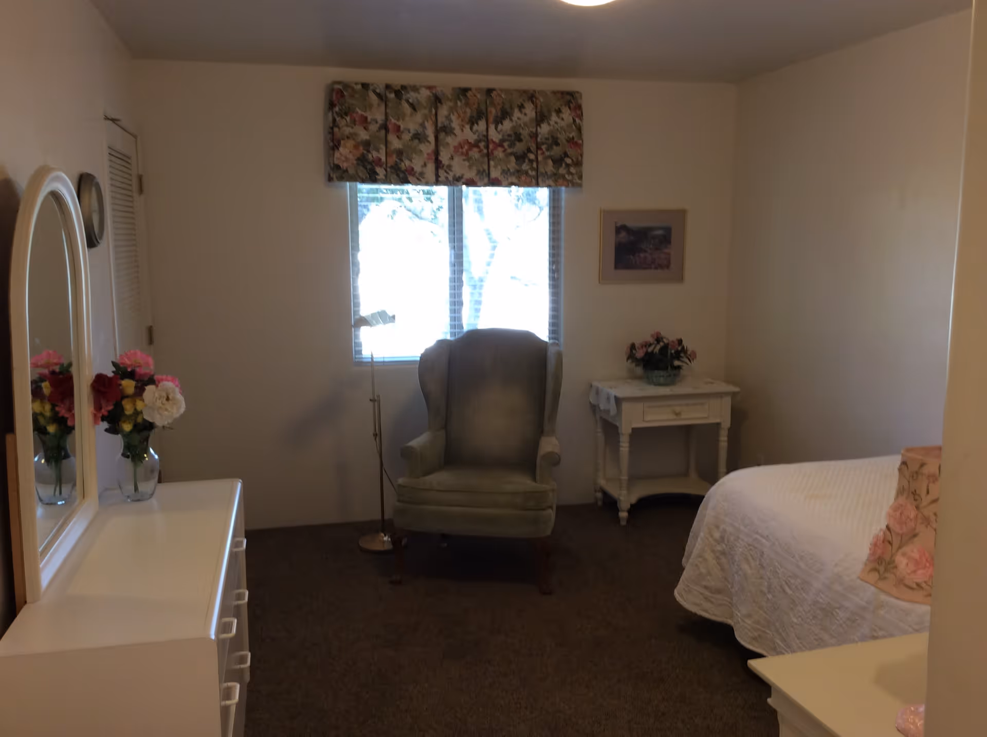 A cozy bedroom with a window covered by floral valance curtains. There is a green upholstered armchair in front of the window, a white nightstand with a flower arrangement, a bed with a white quilt and a floral lampshade, and a white dresser with a mirror and a vase of flowers.