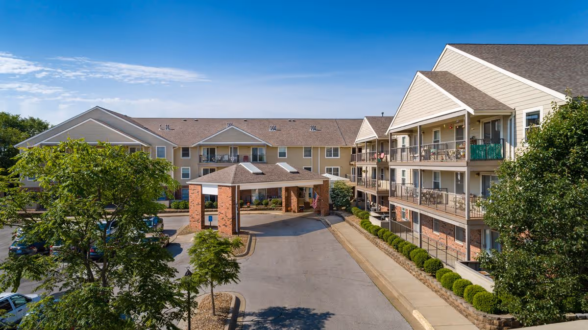 Exterior front entrance of a three-story assisted living building with balconies, a covered porte-cochere, parking area, and surrounding trees.