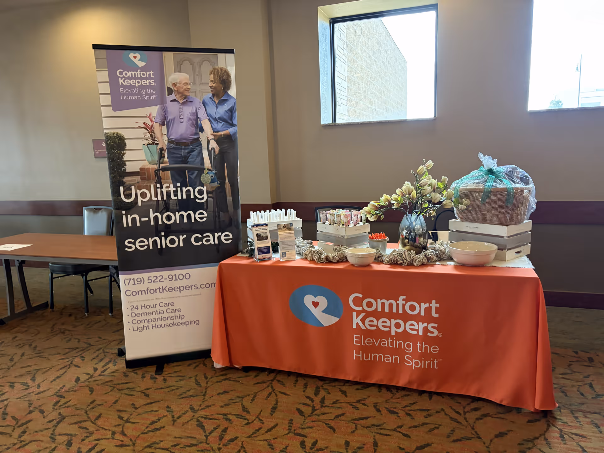 Indoor event setup featuring an orange Comfort Keepers tablecloth and a promotional banner.