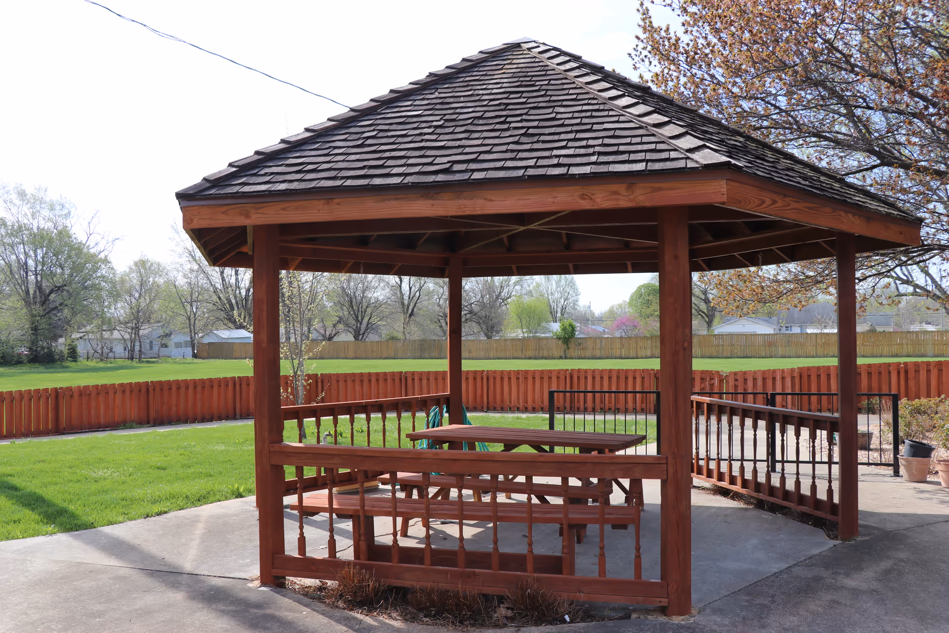 A wooden gazebo with a picnic table sits in a fenced, grassy outdoor area.