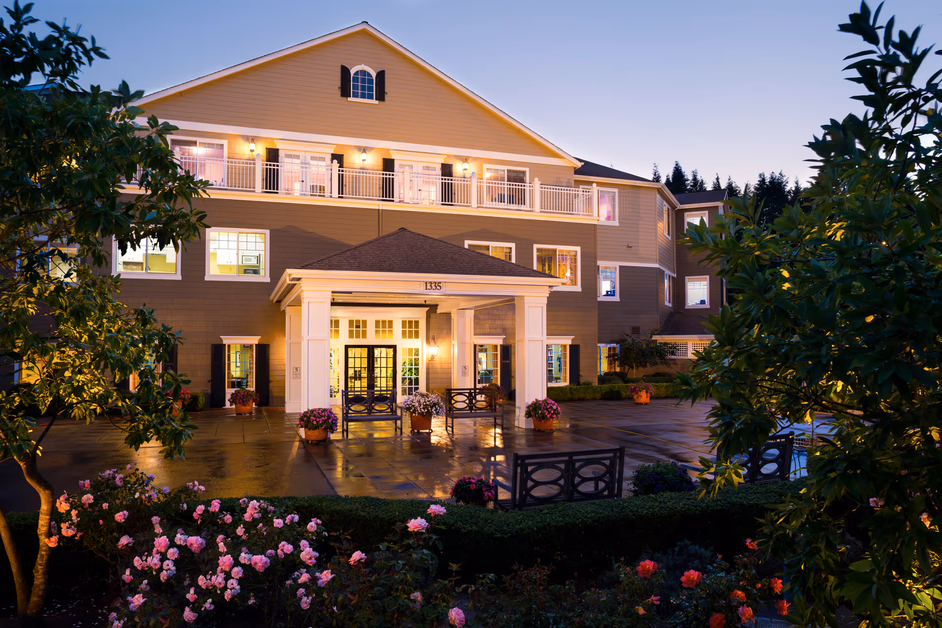Evening exterior view of a three-story senior living building with a lit covered entrance, balconies, and landscaped flowerbeds.