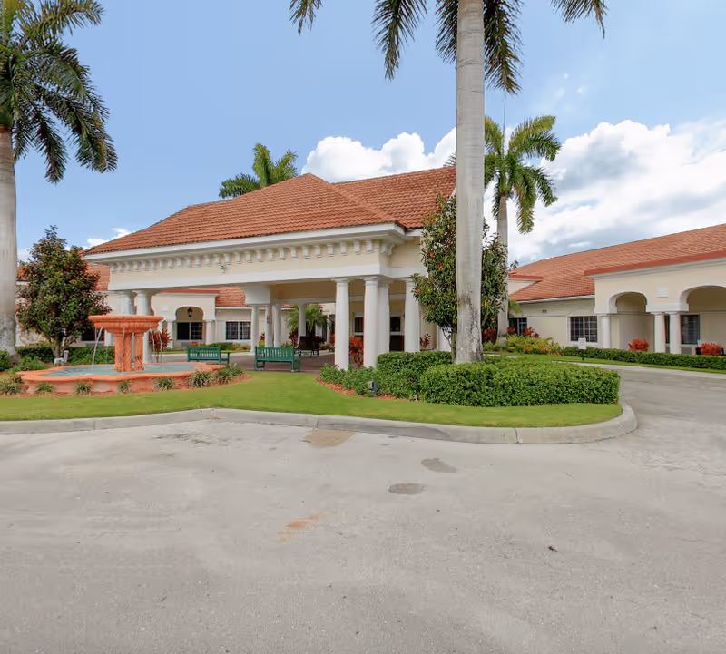 Exterior view of Clearday Living of Naples facility featuring a covered entrance with white columns, a red-tiled roof, palm trees, green benches, and a decorative fountain surrounded by landscaped greenery under a partly cloudy sky.