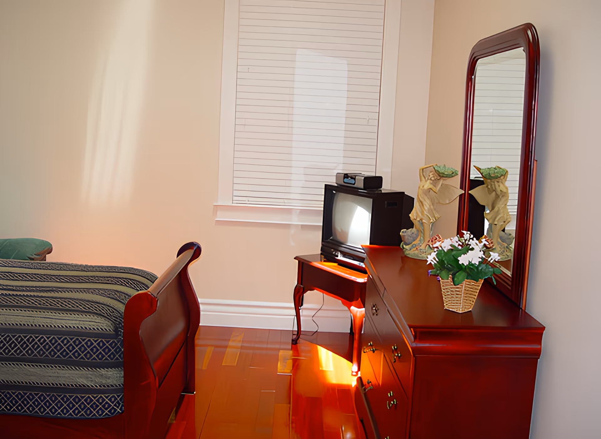 A bedroom with a wooden bed frame partially visible on the left, a wooden dresser with a large mirror on the right, decorated with a small basket of flowers and two angel figurines. A small vintage television sits on a wooden side table in front of a window with closed blinds. The room has polished wooden floors and light-colored walls.