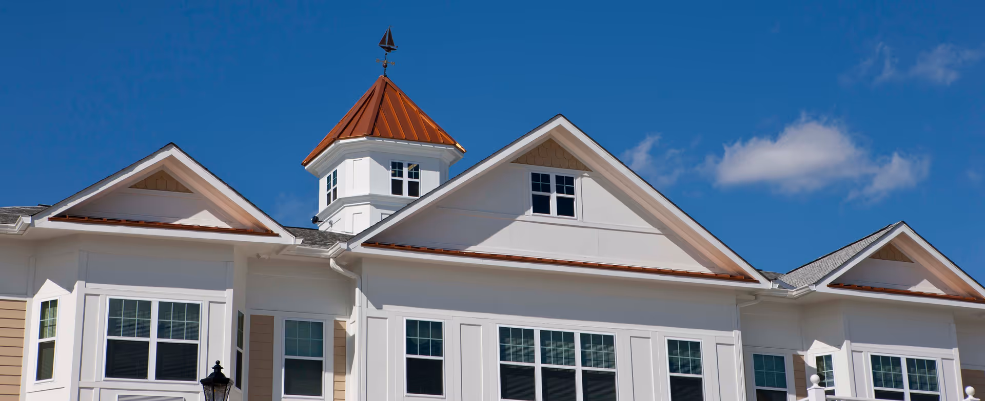 Exterior view of a senior living facility building with white siding, multiple windows, and a small tower with a copper-colored roof under a clear blue sky.