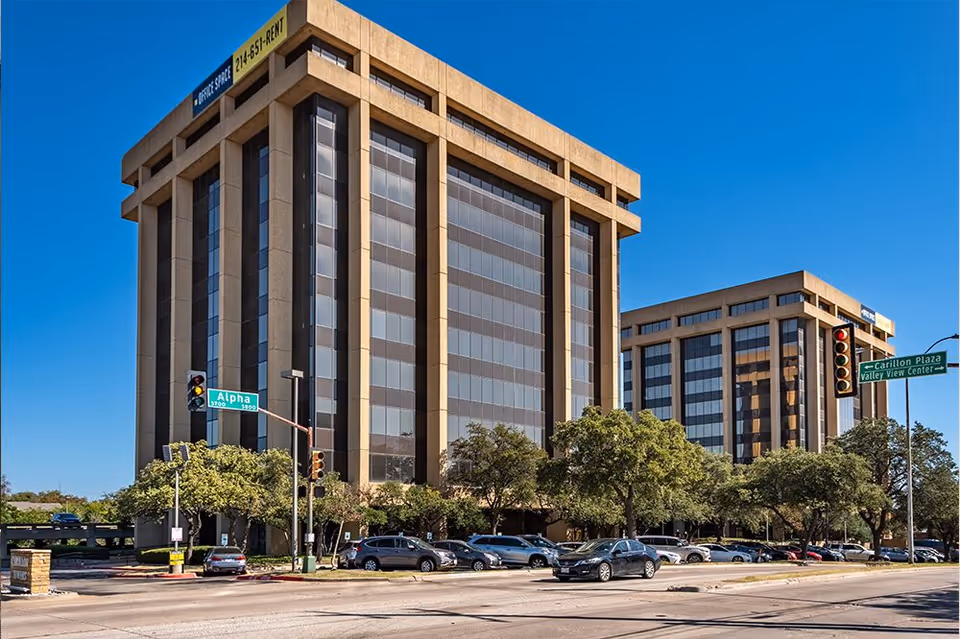 Two tall office buildings along a street with trees, cars, and street signs.