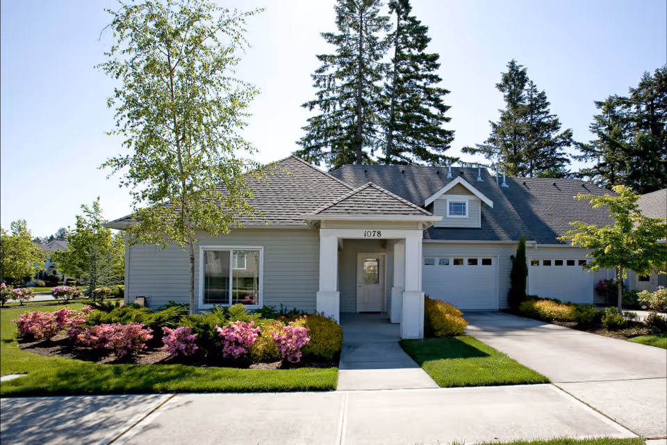 Exterior view of a single-story residential building with a gray roof and light-colored siding. The building has a covered entrance with the number 1078 above the door, a two-car garage, and well-maintained landscaping including green grass, bushes, and flowering plants. Tall trees are visible in the background under a clear blue sky.
