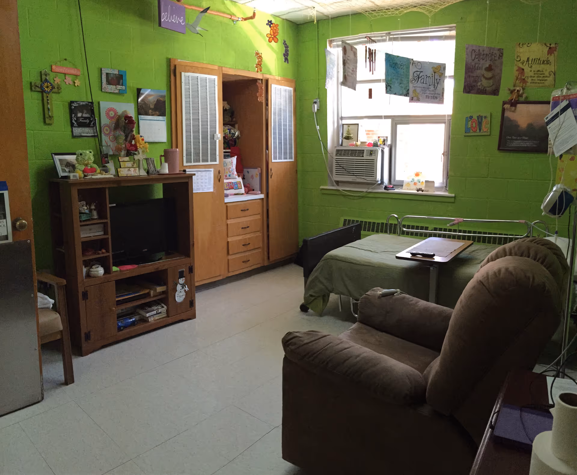 Bright green senior living bedroom with a bed, recliner, cabinet unit, and window air conditioner.