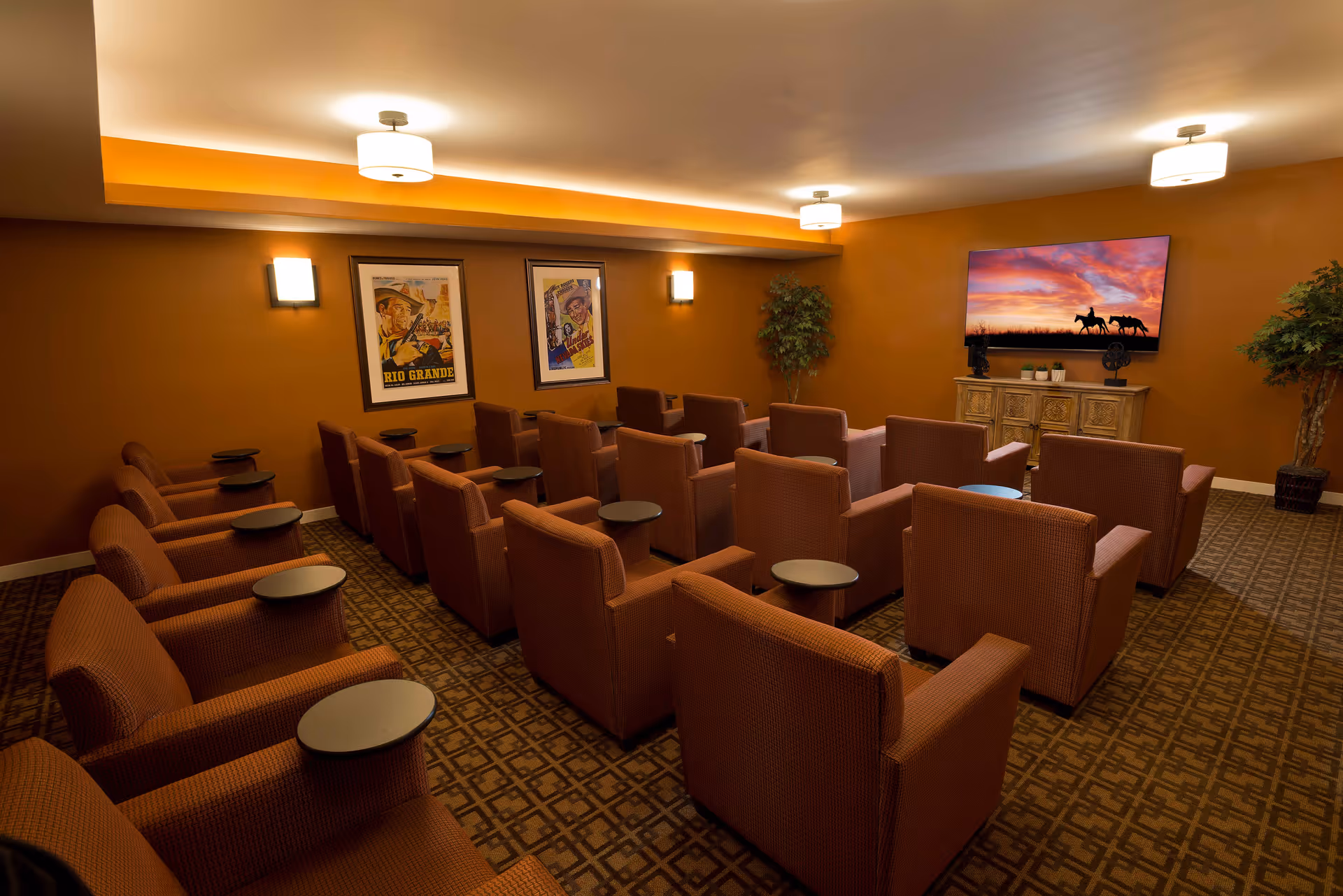 A cozy theater room with multiple rows of brown upholstered armchairs, each equipped with a small round side table. The walls are painted a warm orange color and decorated with two framed vintage movie posters. At the front of the room, a large flat-screen TV displays an image of two people on horseback against a colorful sunset sky. There are two potted plants placed in the corners of the room, and the ceiling has recessed lighting with three round light fixtures.