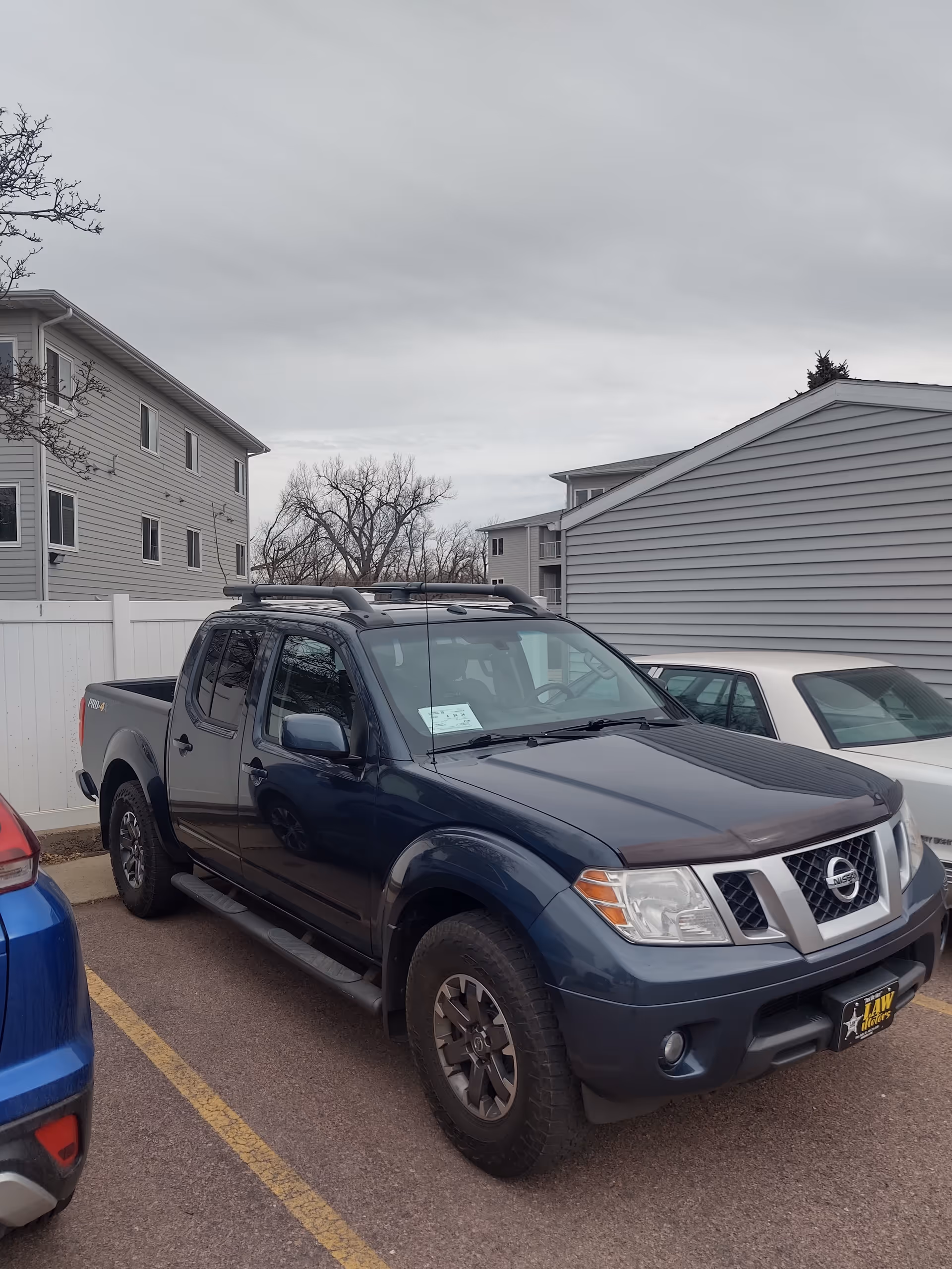 Blue Nissan pickup parked in a lot beside apartment buildings under cloudy sky.