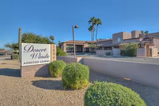 Exterior view of Desert Winds Assisted Living facility showing the main building, desert landscaping with bushes and palm trees, and a sign with the facility name and address number 2054.