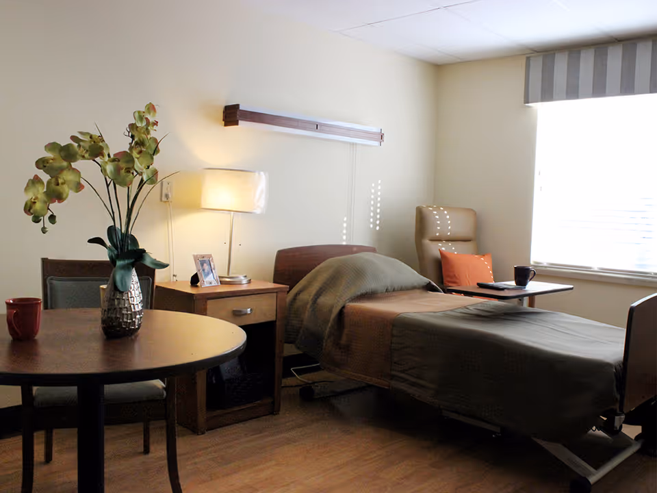 A cozy bedroom in a healthcare facility featuring a single bed with a green blanket and brown sheets, a bedside table with a lamp and a framed photo, a round table with a vase of flowers and a red mug, and a comfortable chair with an orange pillow near a window with blinds.