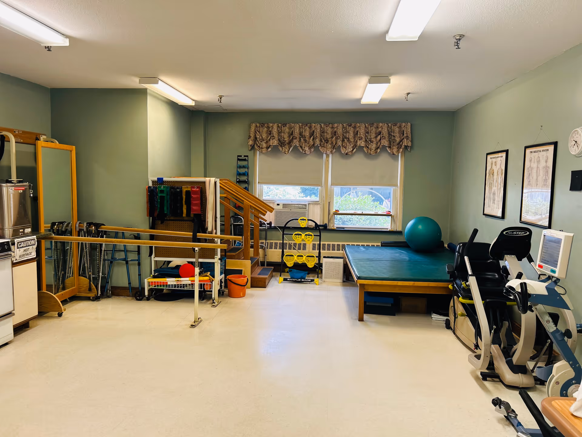 A physical therapy room with exercise equipment including parallel bars, a recumbent exercise bike, a large exercise ball, a padded therapy table, and various therapy tools. The room has green walls, two windows with floral valances, and anatomical charts on the wall.