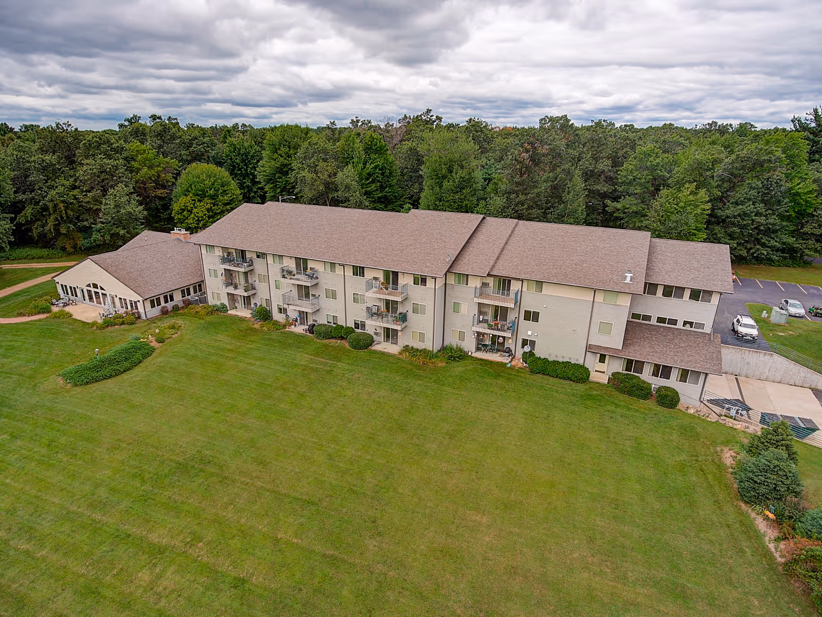 Aerial view of a multi-story senior living facility named MasonWoods surrounded by a large green lawn and trees under a cloudy sky. The building has balconies and a parking area with a few cars visible.