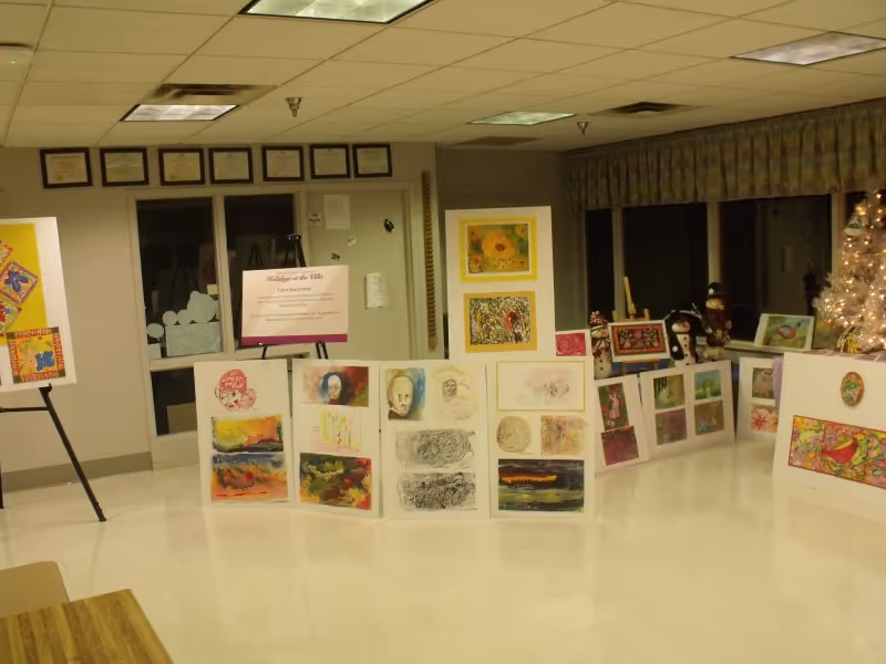 Interior community room with easels and folding panels displaying colorful artwork and framed certificates on the wall.