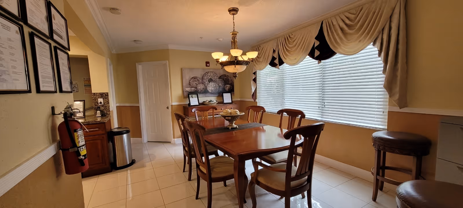 A dining room with a wooden table, chairs, chandelier, and large sunlit windows.