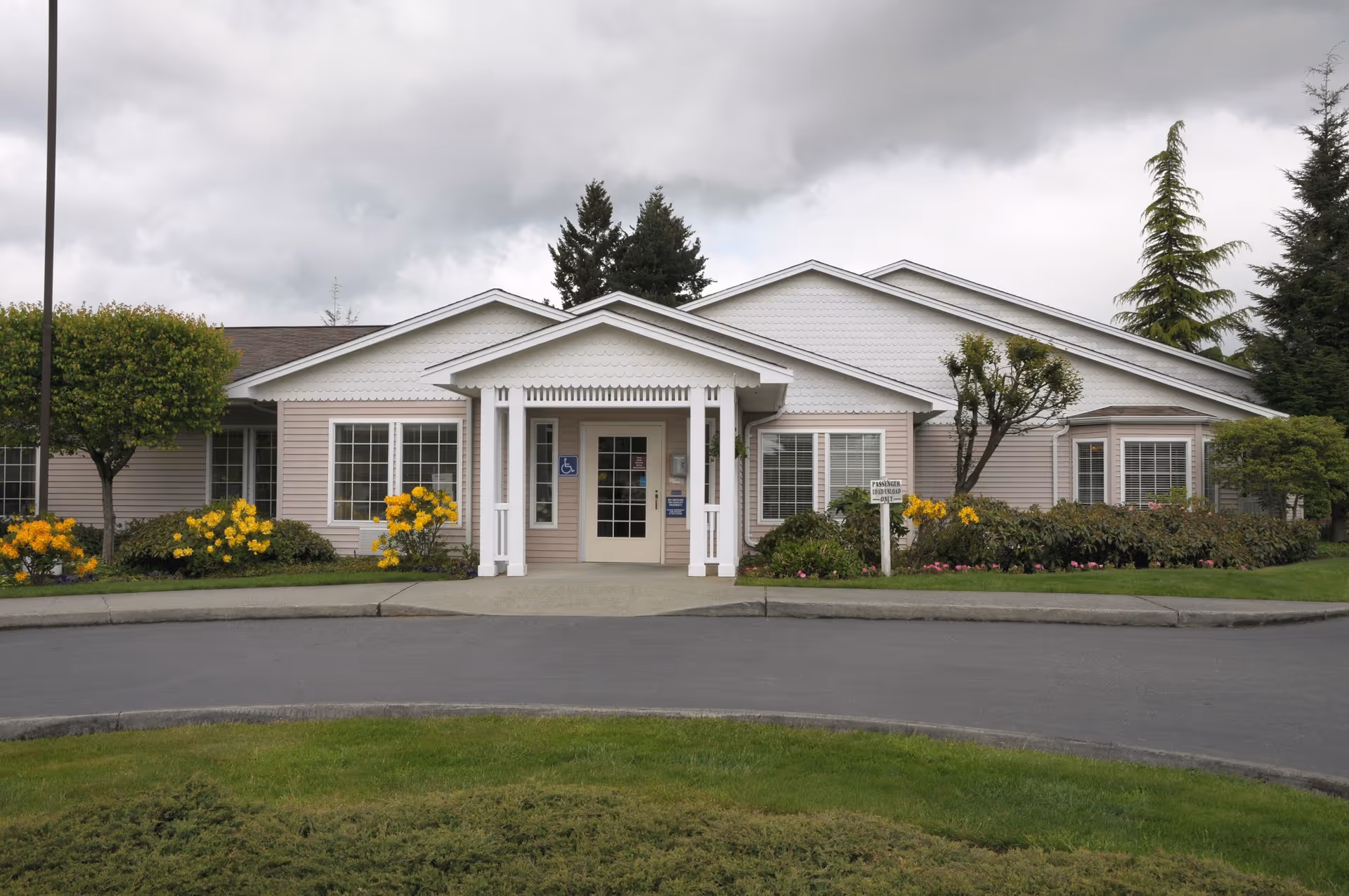 Front entrance of a single-story care facility with a covered portico, landscaped flowerbeds, and a circular driveway.