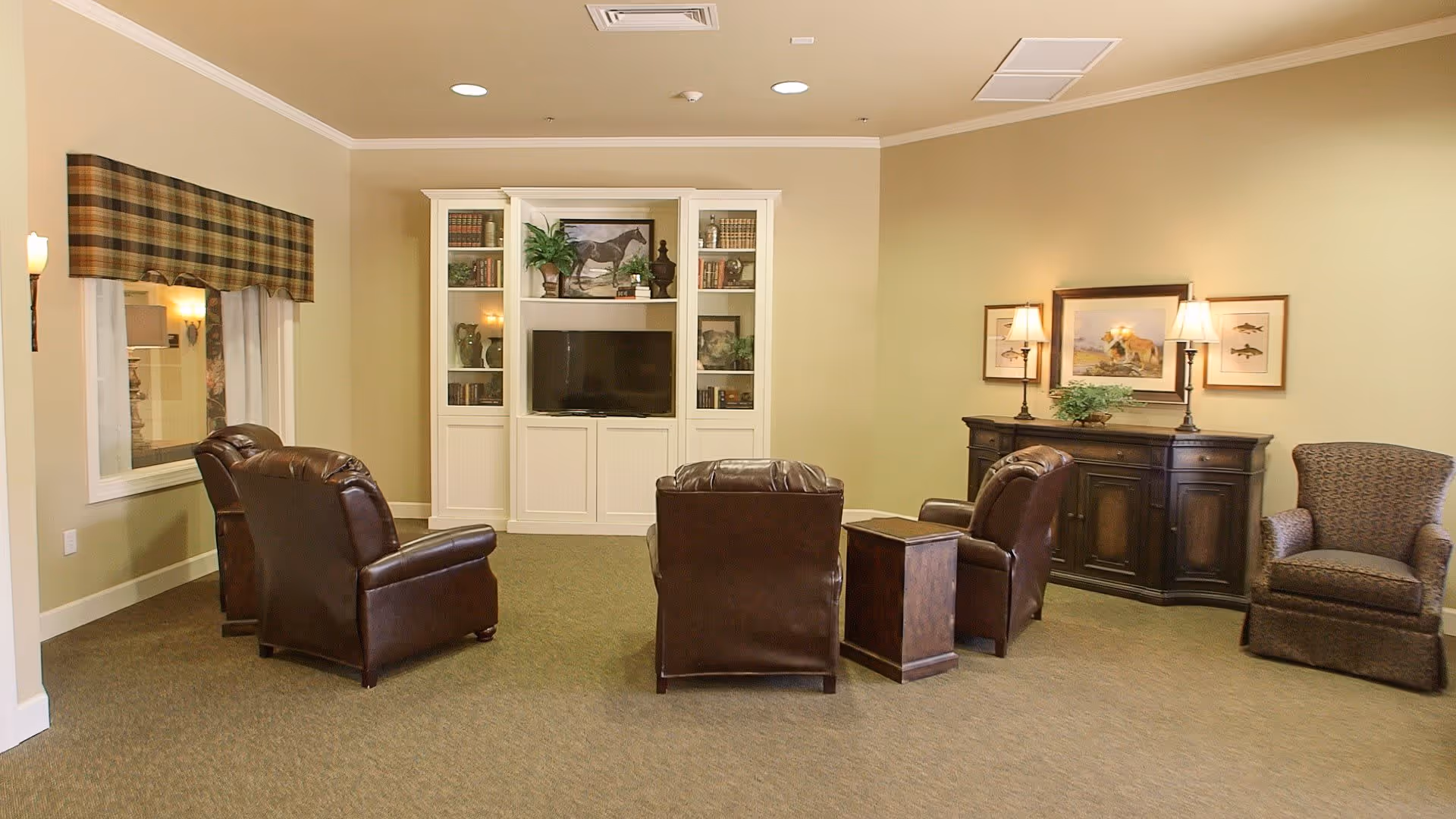 A cozy senior living common area with four brown leather armchairs arranged around a small wooden table, facing a white built-in entertainment center with a TV and decorative items. The room has beige walls, carpeted floor, a window with a plaid valance, and a dark wooden sideboard with two lamps and framed artwork above it.
