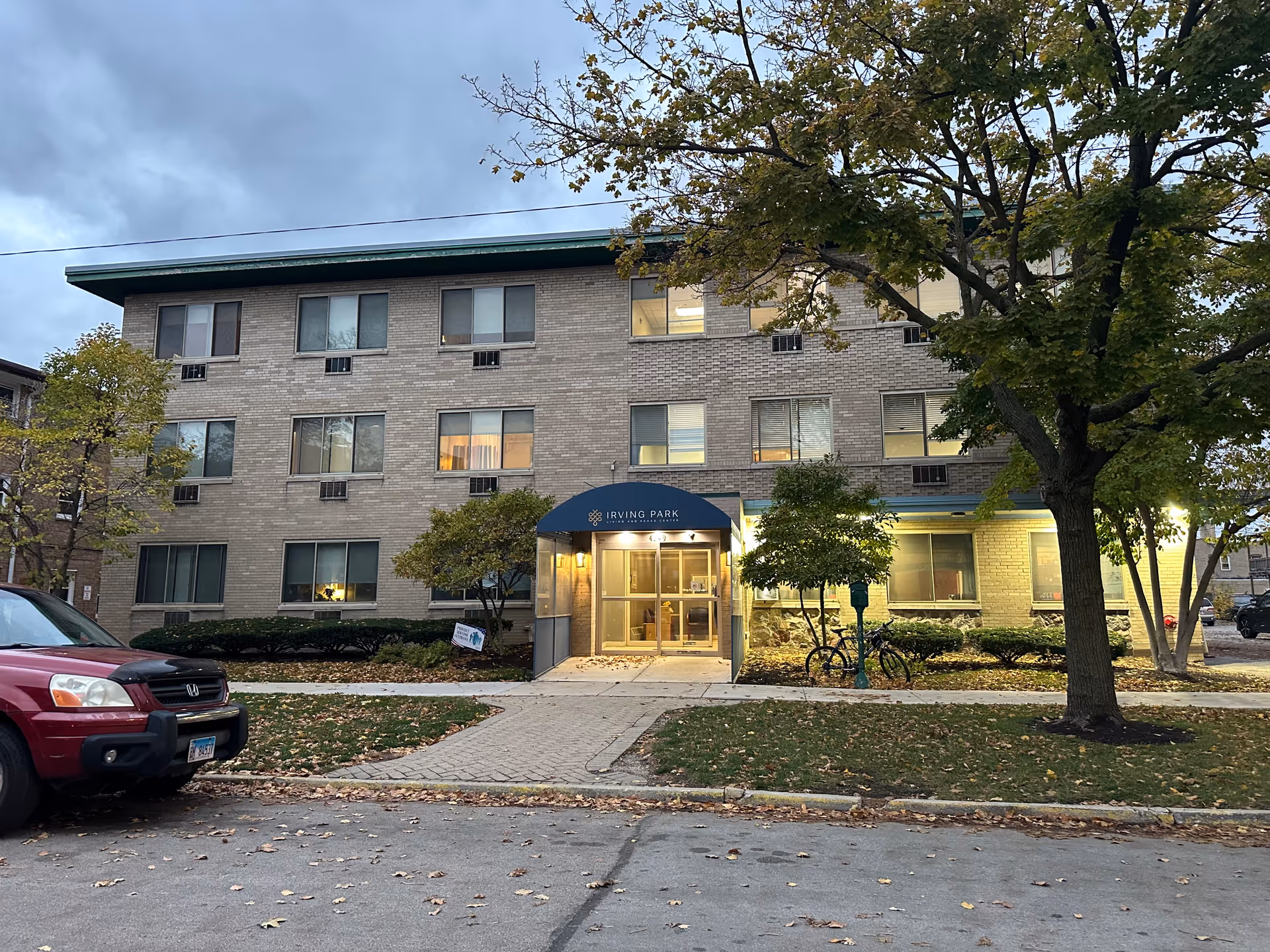 Exterior view of a three-story brick building with a blue awning above the entrance that reads 'Irving Park'. The building is surrounded by trees with autumn leaves and a red SUV is parked on the street in front. The sky is cloudy and it appears to be early evening.