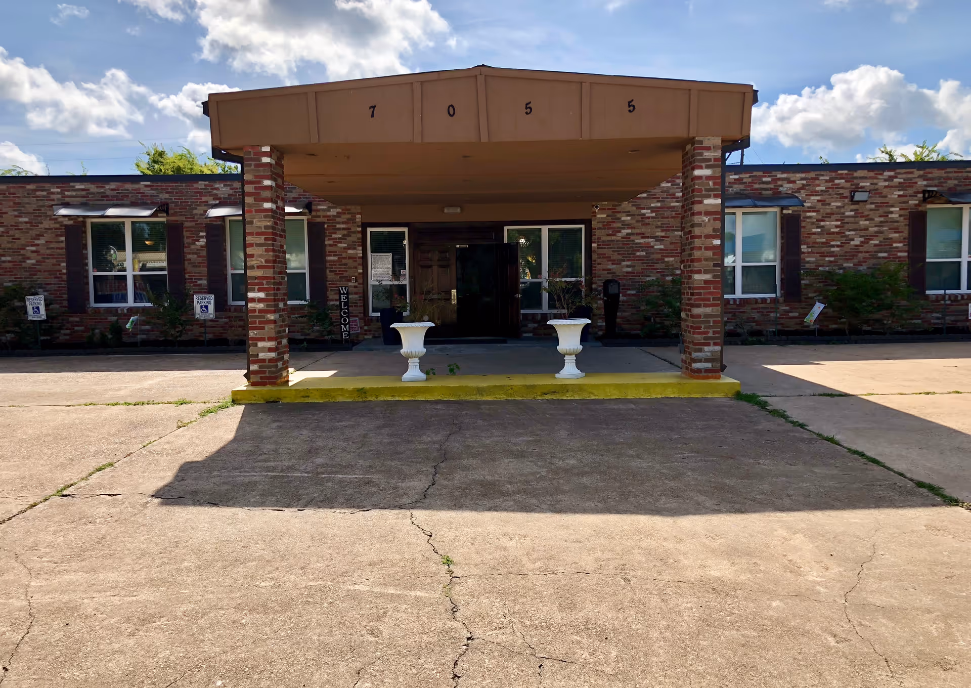 Front entrance of a brick building with a covered porch supported by two brick columns. The building has several windows with white frames and a sign that says 'WELCOME' near the door. The address number 7055 is displayed on the porch roof. There are two white planters on the porch and a concrete driveway in front.