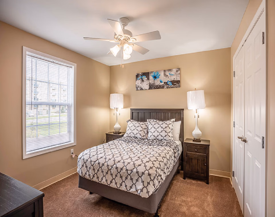 A cozy bedroom with a bed, two nightstands, lamps, ceiling fan, and window.