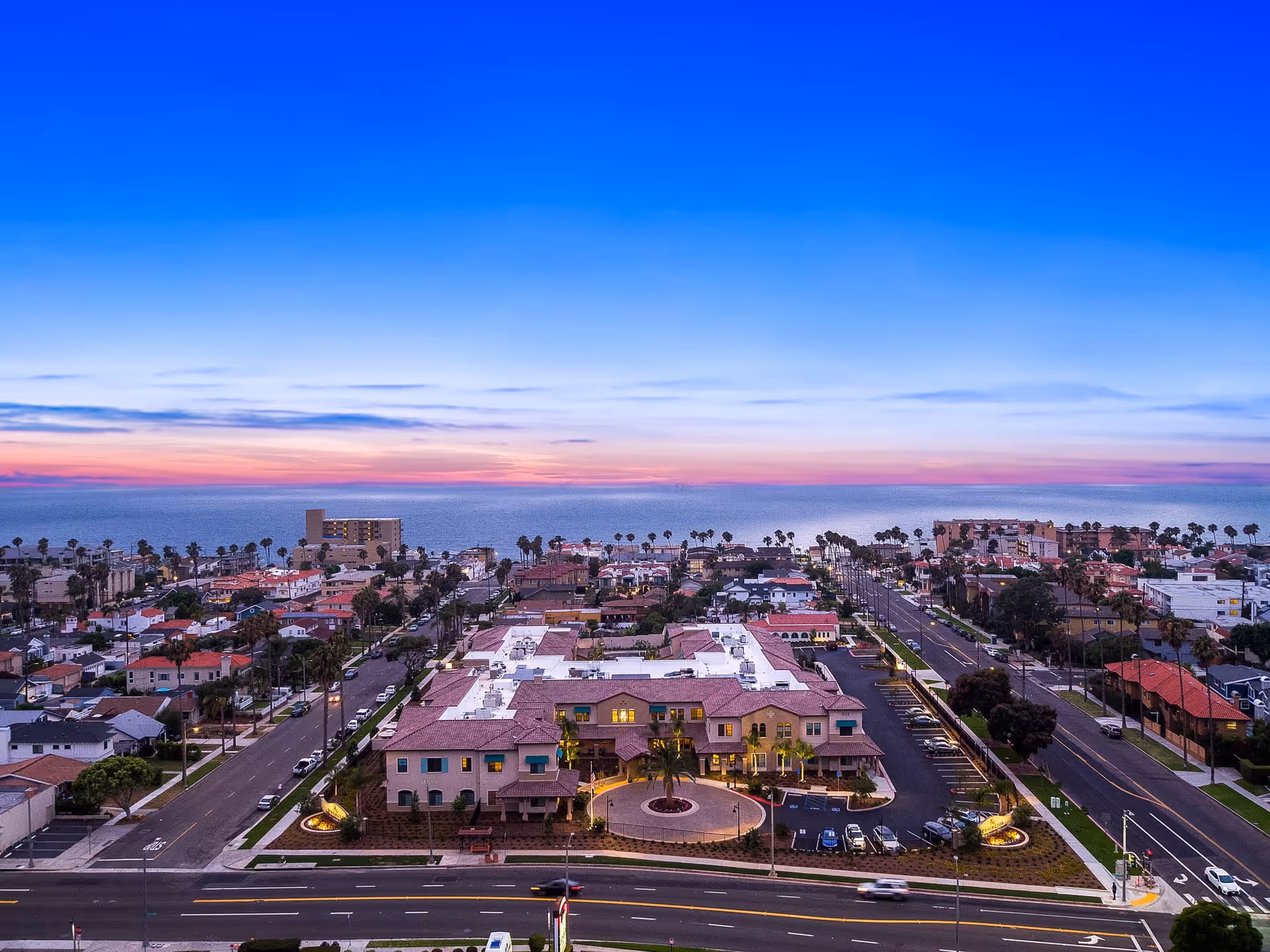 Aerial view of a coastal senior living complex and surrounding neighborhood at sunset with the ocean on the horizon.