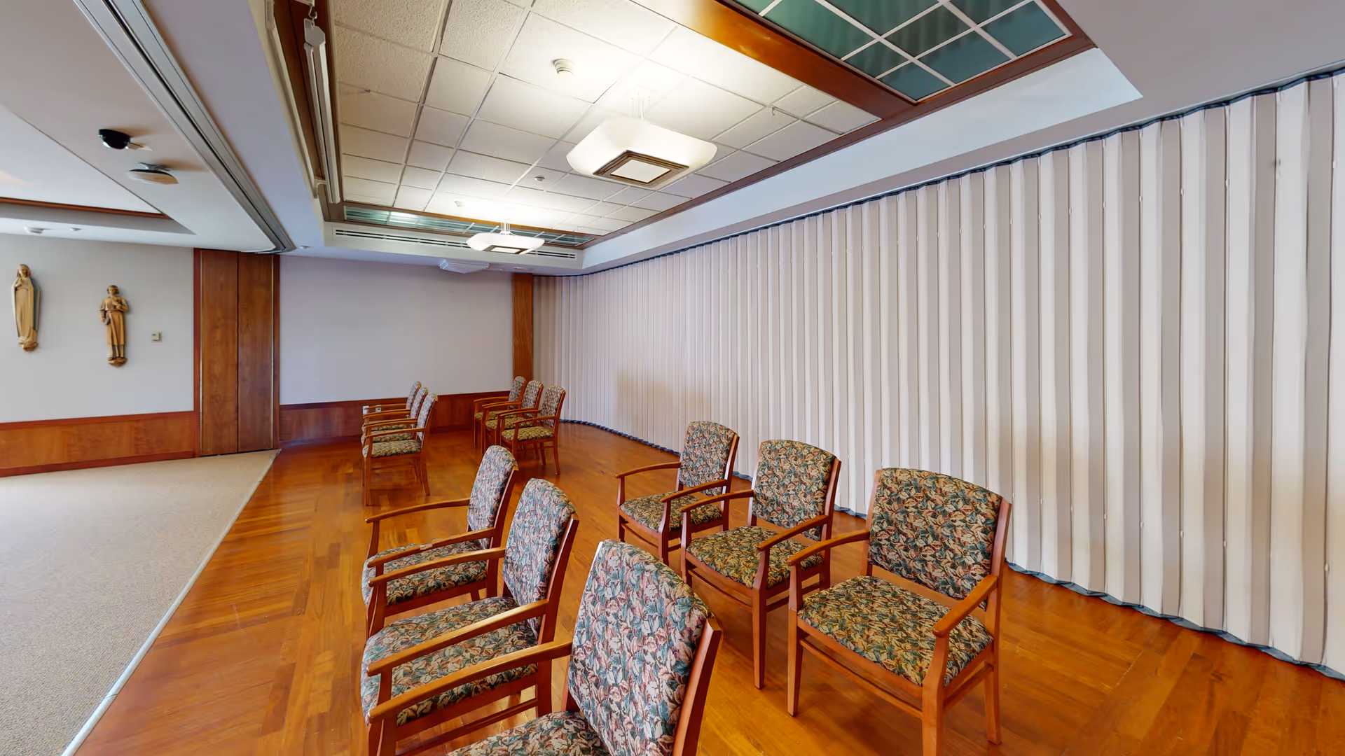 Interior meeting room with wooden floor, patterned chairs, and religious statues along the wall.