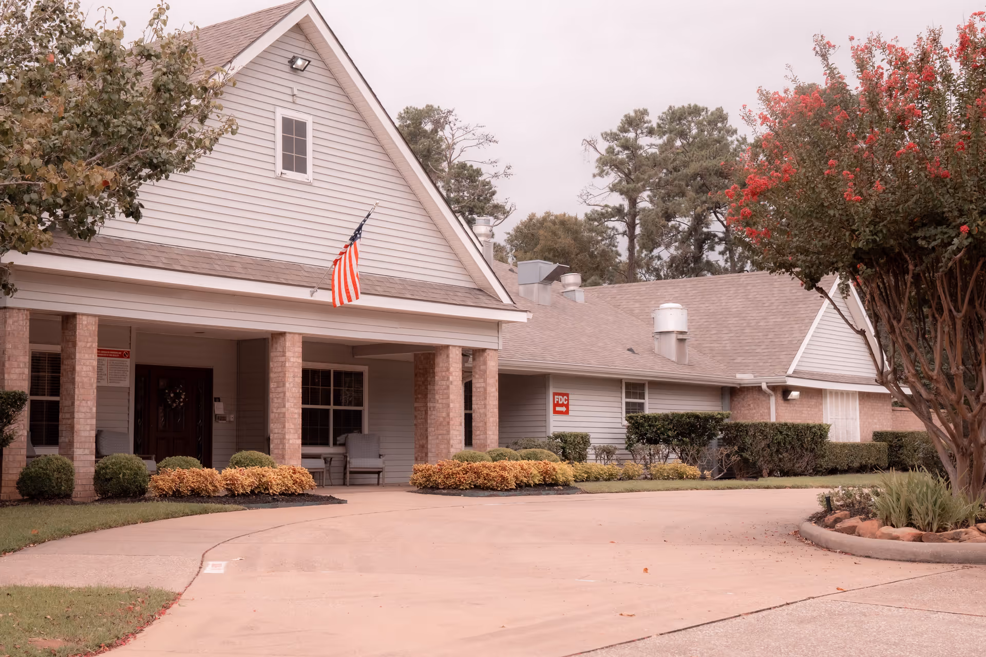 Exterior view of a single-story building with a covered entrance supported by brick columns. The building has light gray siding and a gabled roof. An American flag is mounted near the entrance. There are neatly trimmed bushes and landscaping around the building, with trees on either side. The driveway curves in front of the entrance.