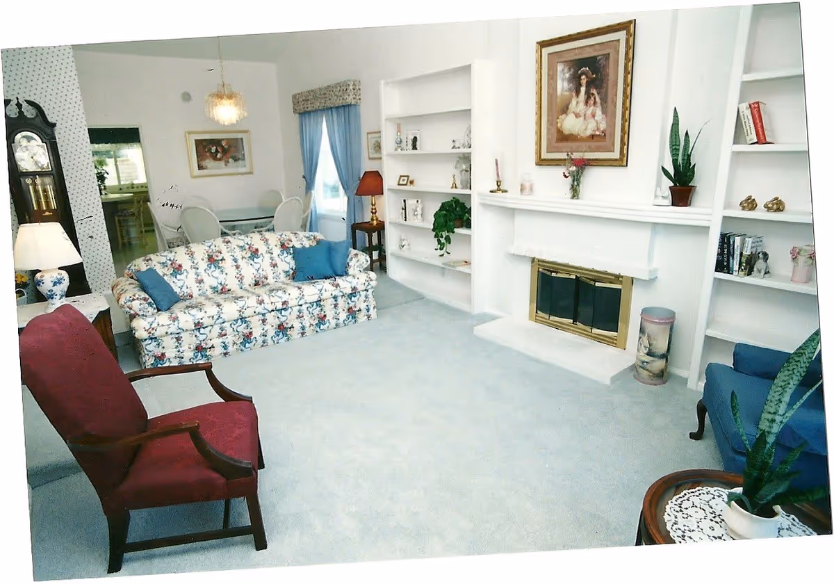 A cozy living room with a floral patterned sofa with blue cushions, a red armchair, a blue armchair, and a fireplace with a gold-trimmed screen. The room has built-in white shelves with decorative items and plants, a framed painting above the fireplace, a grandfather clock, and a dining area with a glass table and white chairs in the background.