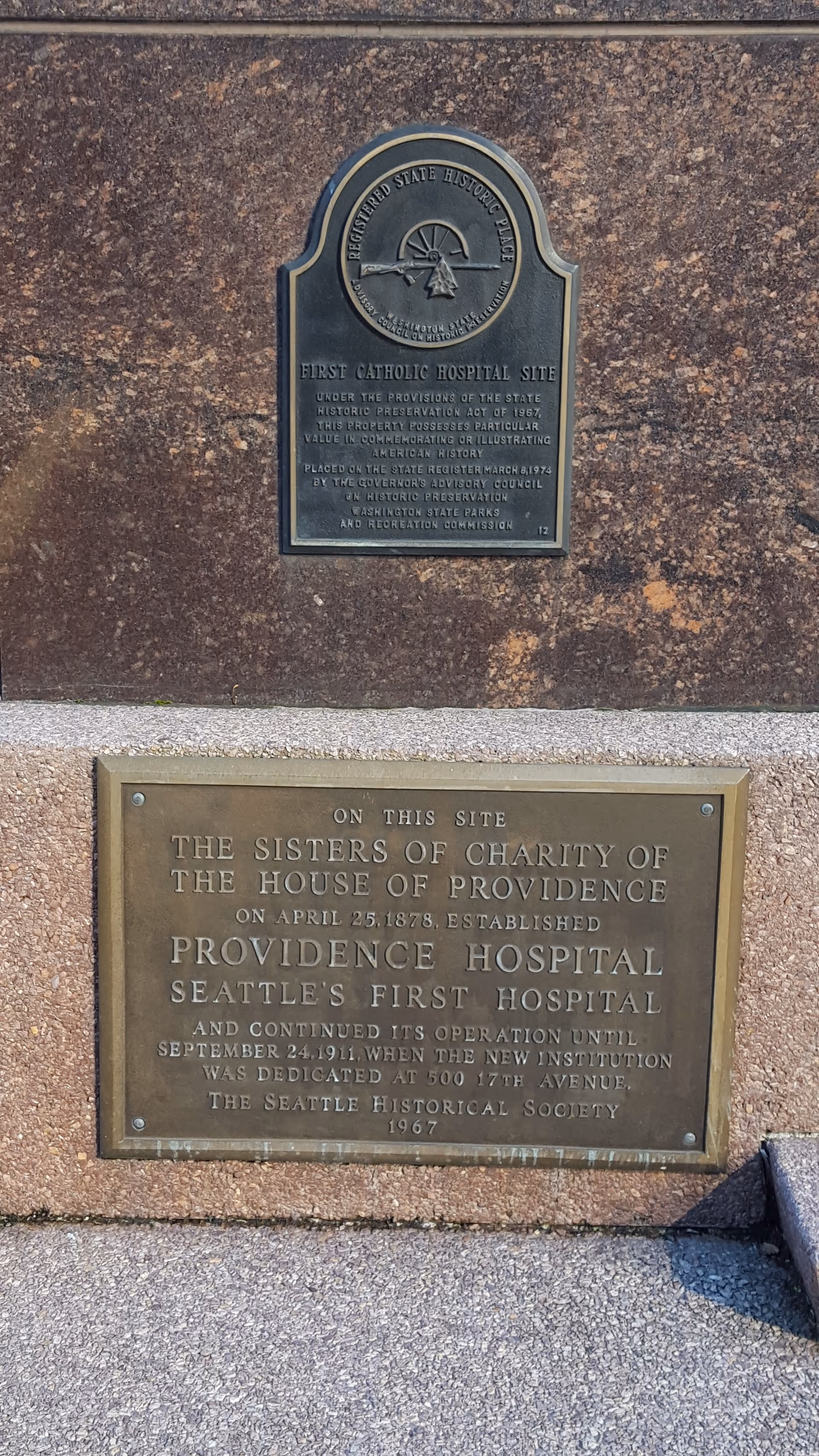 Two bronze plaques mounted on a brown stone exterior wall.