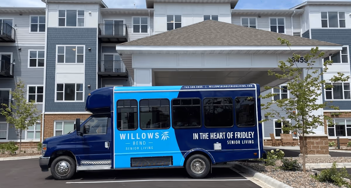 A blue Willows Bend Senior Living shuttle parked in front of the entrance to a multi-story senior living building.