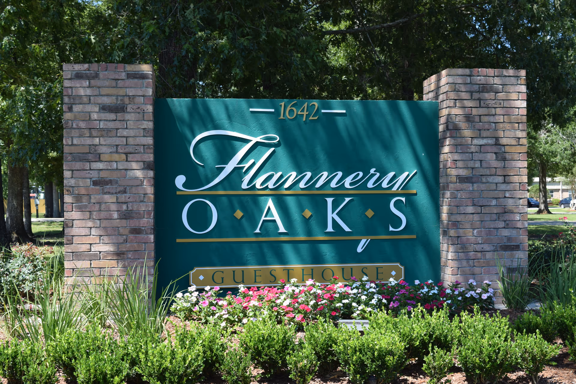 A green sign with gold and white lettering that reads '1642 Flannery Oaks Guest House' set between two brick pillars, surrounded by green bushes and colorful flowers with trees in the background.