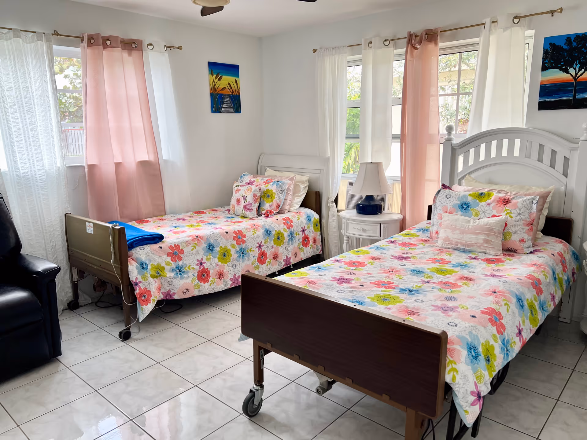 Two floral-patterned hospital beds with white furniture and pink and white curtains in a bright bedroom.