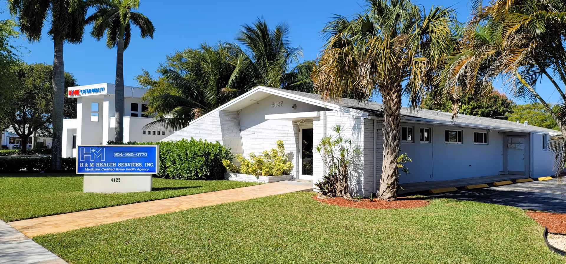 A single-story white health services facility with a blue sign and palm trees at the entrance.