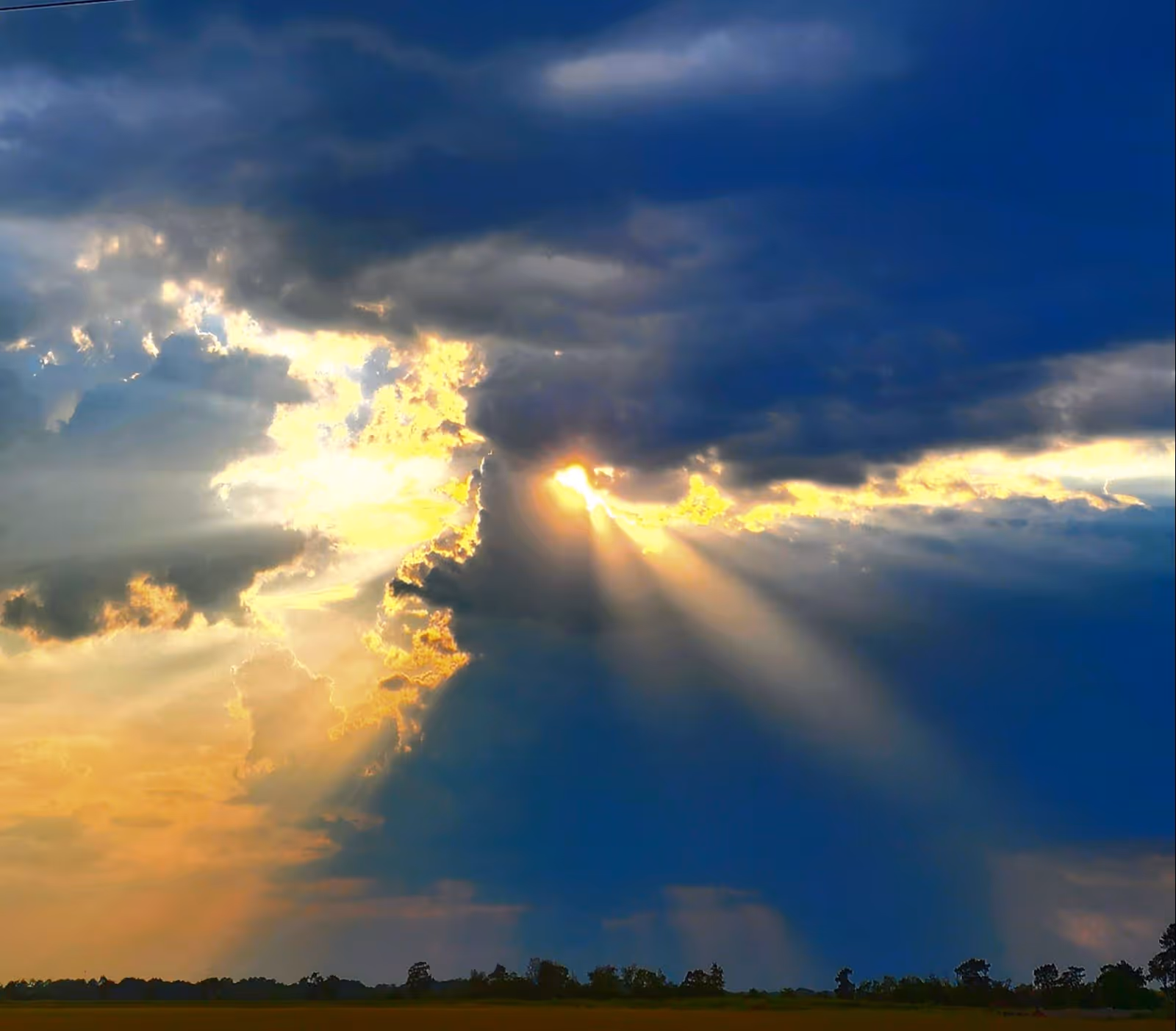 Golden sunbeams break through dark clouds over a flat field with trees.