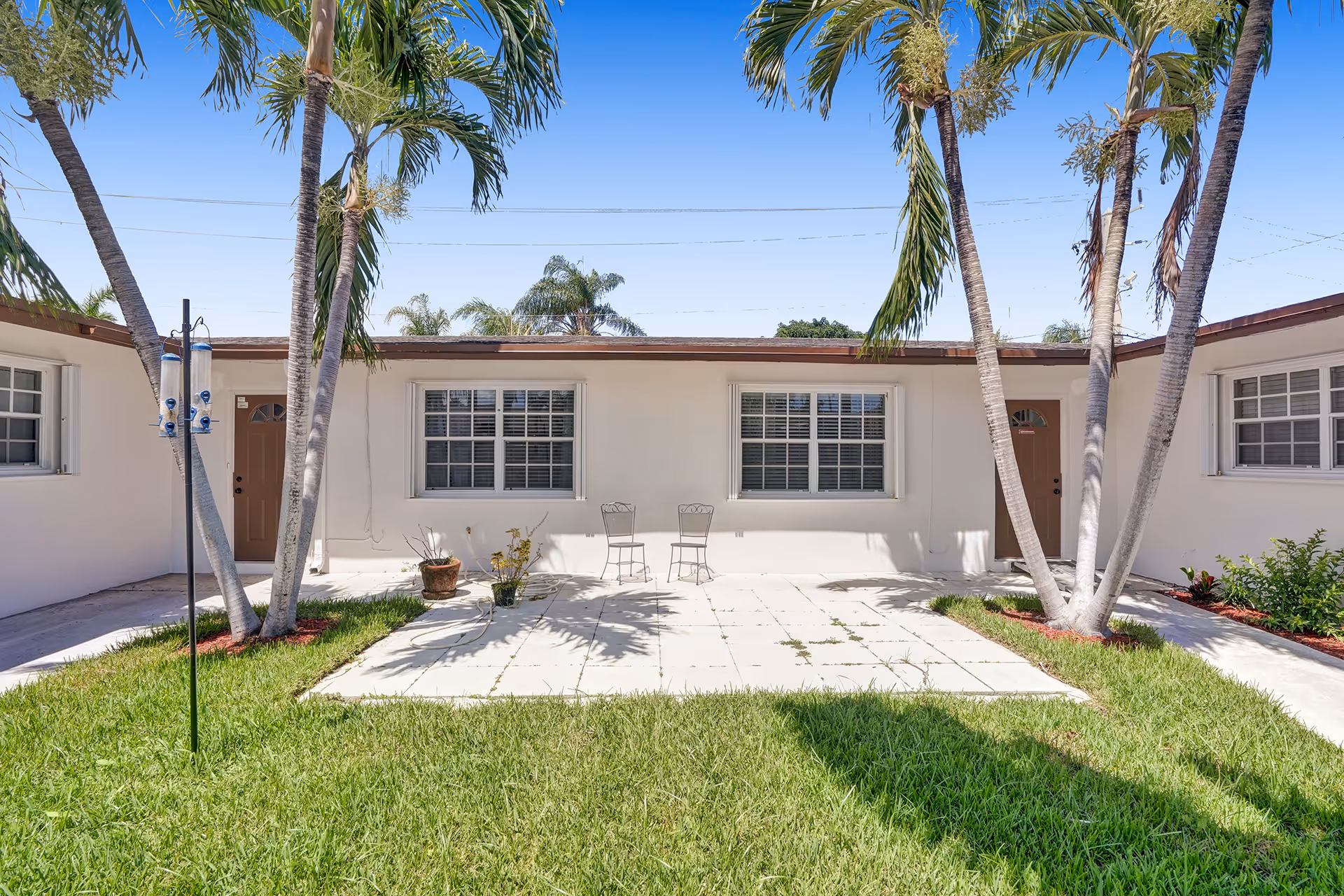 White, single-story facility with a paved courtyard, palm trees, and two metal chairs.