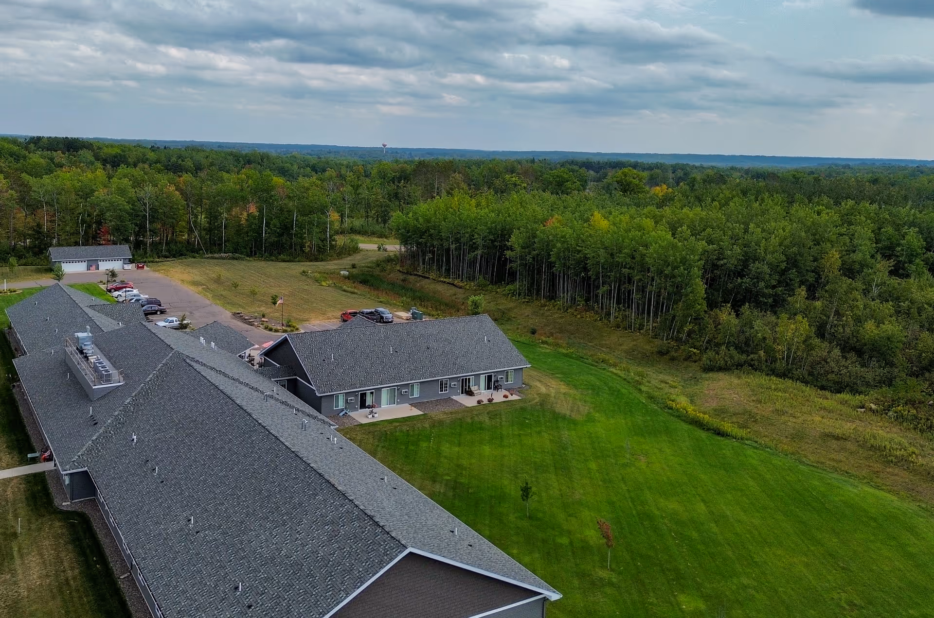 Aerial view of a senior living facility named Golden Horizons Of Sandstone surrounded by green lawns and dense forest. The facility consists of long, single-story buildings with gray roofs and a parking area with several cars. The sky is partly cloudy.