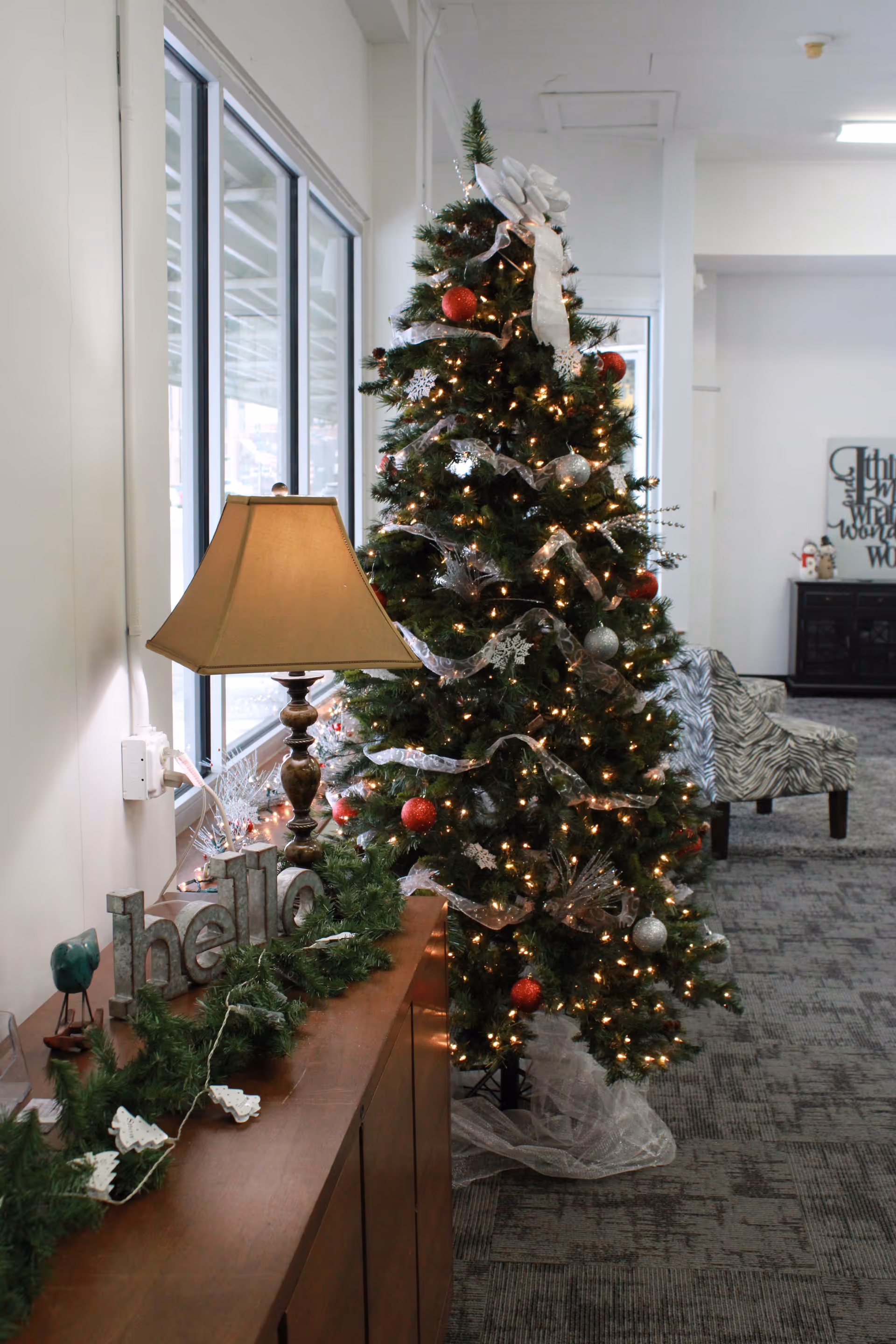 Festively decorated indoor lobby with a Christmas tree, garlands, lamp, and seating.