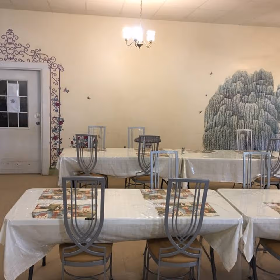 A dining room with several tables covered in white tablecloths and placemats. Each table is surrounded by metal chairs with unique backrest designs. The walls are decorated with a mural of a large tree and floral patterns near a door. A chandelier with three lights hangs from the ceiling.