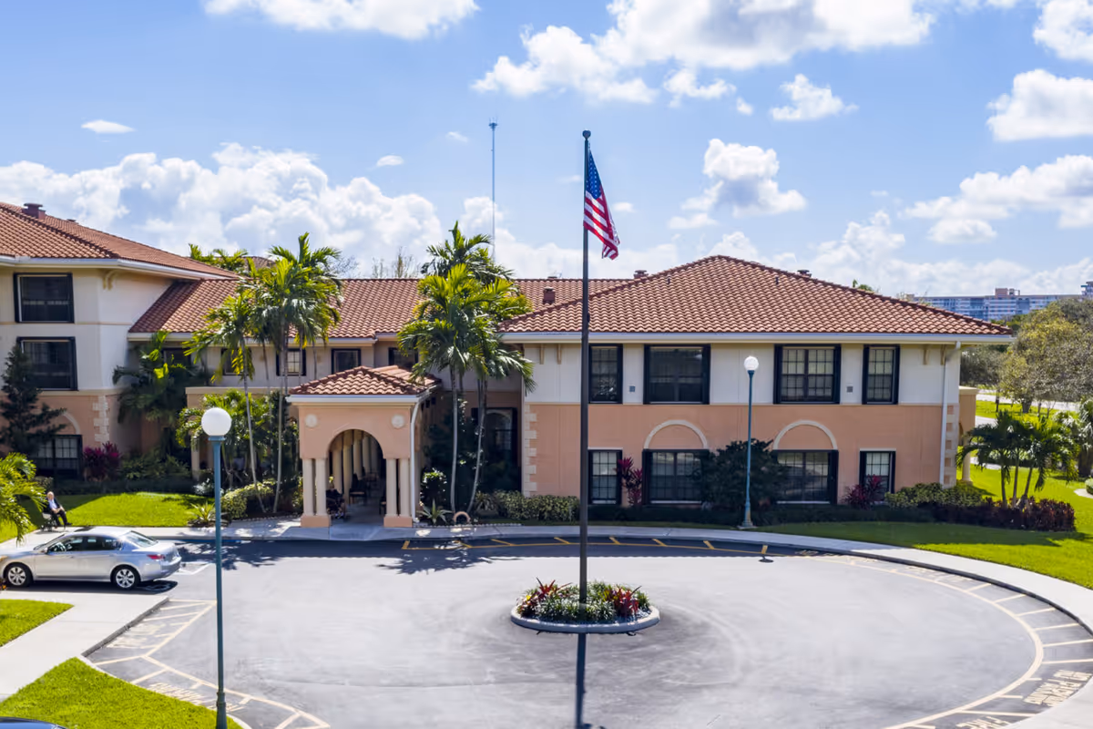 A senior living facility building with a circular driveway, flagpole, and palm trees.