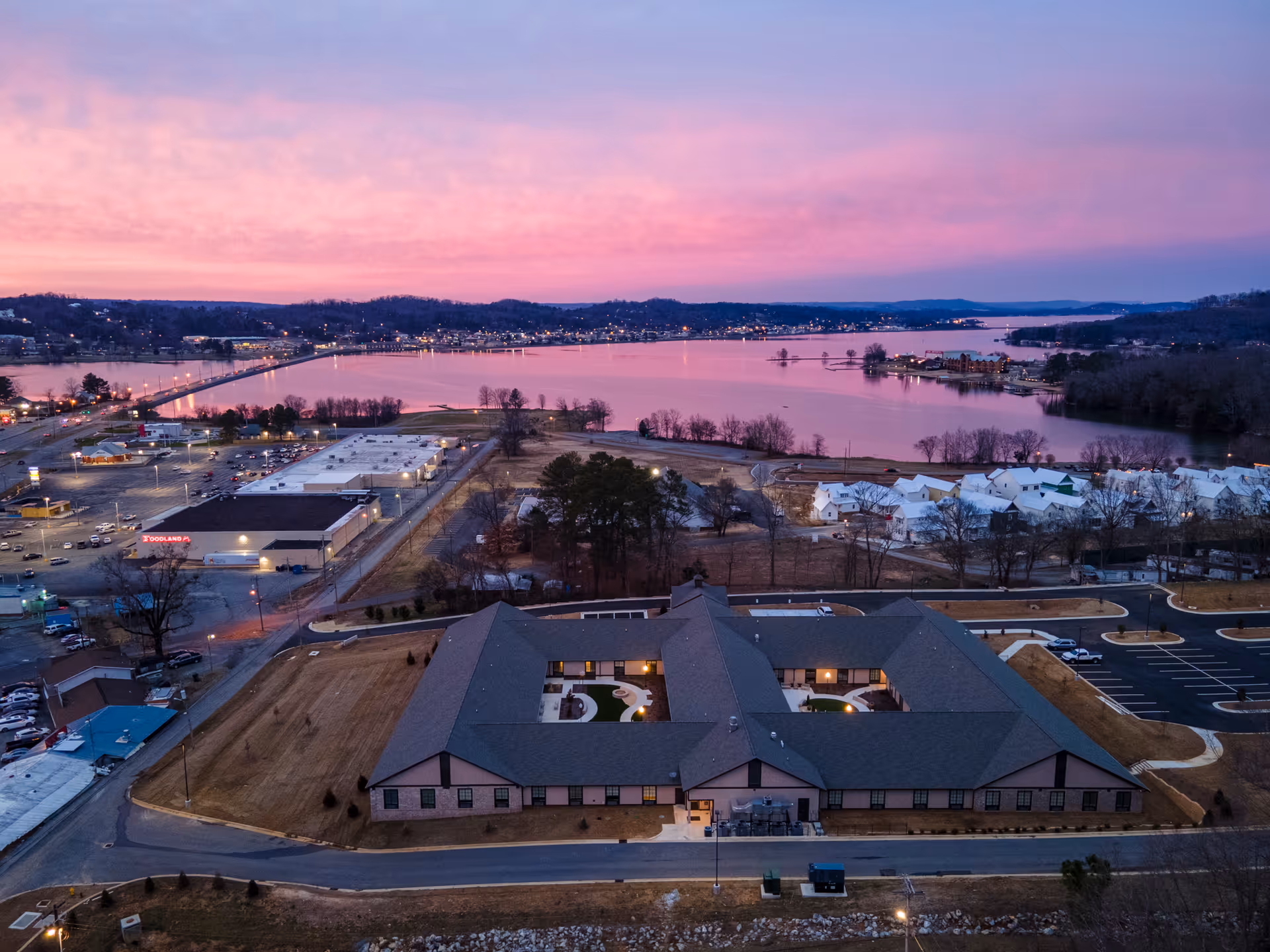 Aerial view of a lakeside senior living campus at sunset, with a U-shaped building and parking.