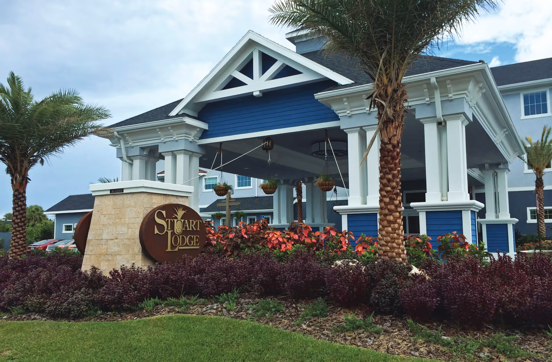 Covered entrance of a blue-and-white lodge with palm trees, landscaped flowerbeds, and a 'Stuart Lodge' sign.