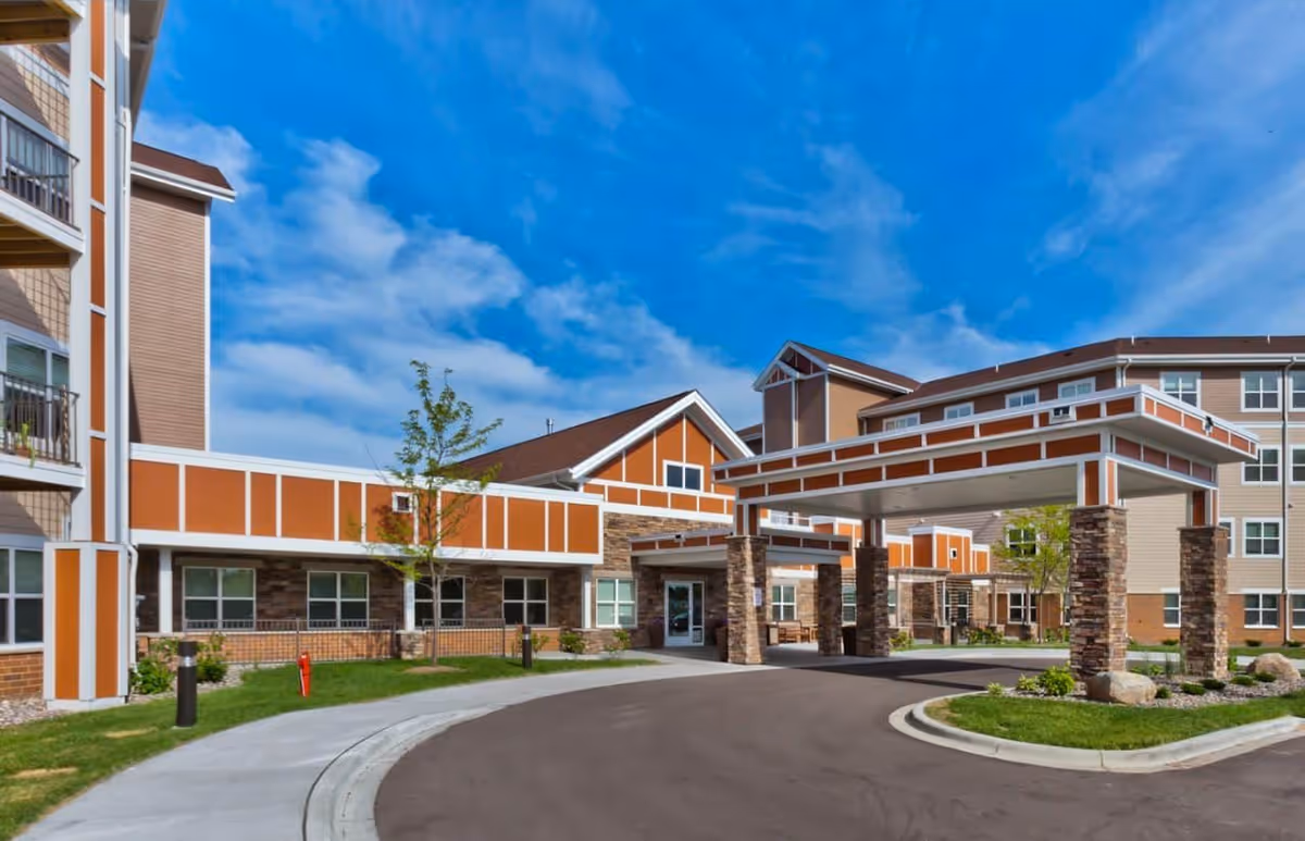 Front entrance of a multi-story senior living building with a covered porte-cochere, stone columns, and orange-beige siding under a blue sky.