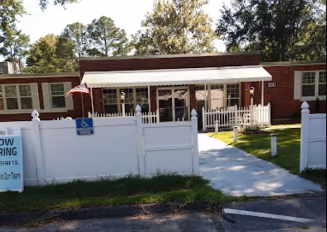 Single-story brick building entrance with a white picket fence, concrete walkway under a metal awning, and an American flag.