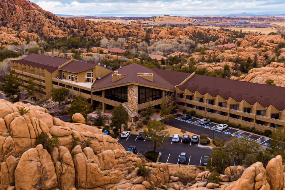 Aerial view of Granite Gate Senior Living facility surrounded by large rock formations and desert landscape. The building has a brown roof and beige exterior with multiple windows and a parking lot with several cars parked. Trees and shrubs are scattered around the property.